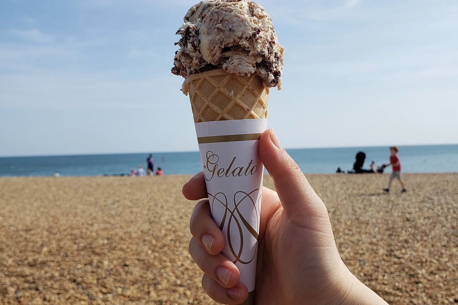 A scoop of gelato in a waffle cone, held up on a beach.