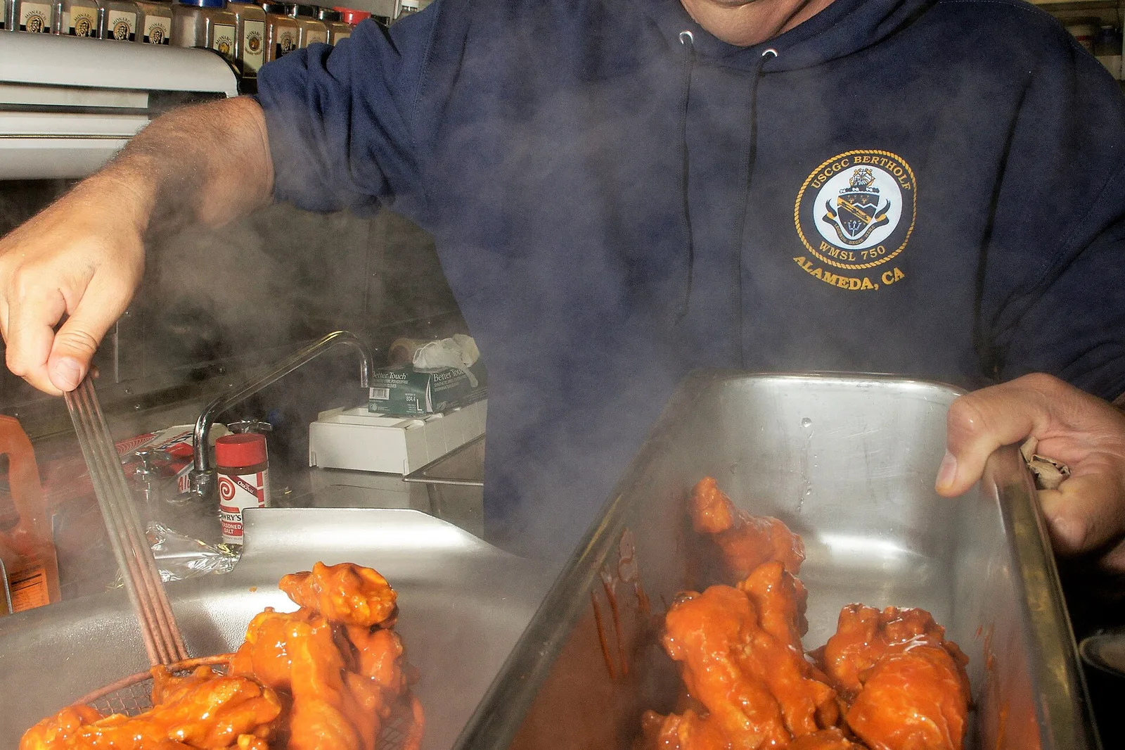 A cook tosses buffalo chicken wings in sauce in a ship's galley.